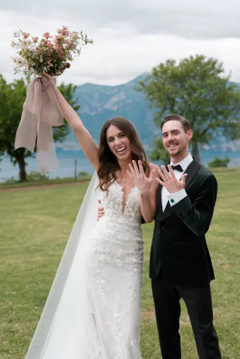Bride and groom celebrating with their wedding rings and bouquet against lake and mountain views at Rustico Bertel.
