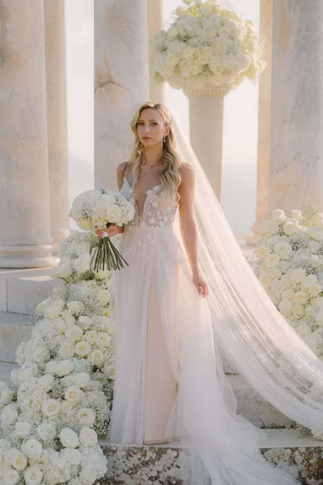 Bride in a flowing gown with a long veil holding a white bouquet among lush floral arrangements at Son Marroig’s pavilion.