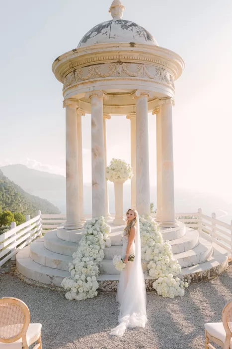 Bride standing with bouquet among cascading white flowers at Son Marroig’s pavilion, veil trailing behind her.