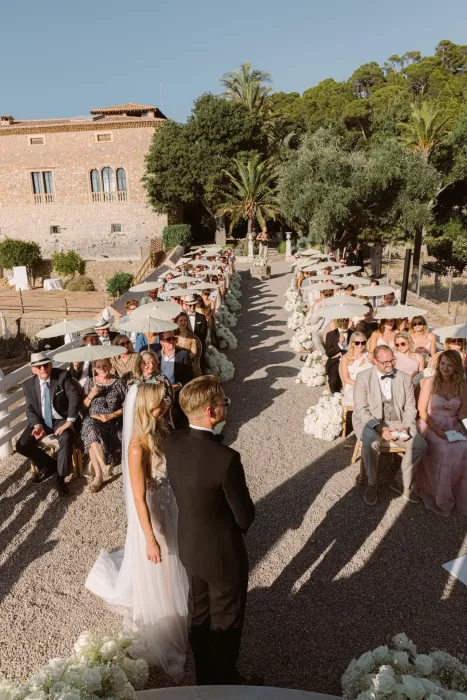 Wedding guests seated with parasols lining the floral aisle at Son Marroig as the bride and groom stand together at the altar.