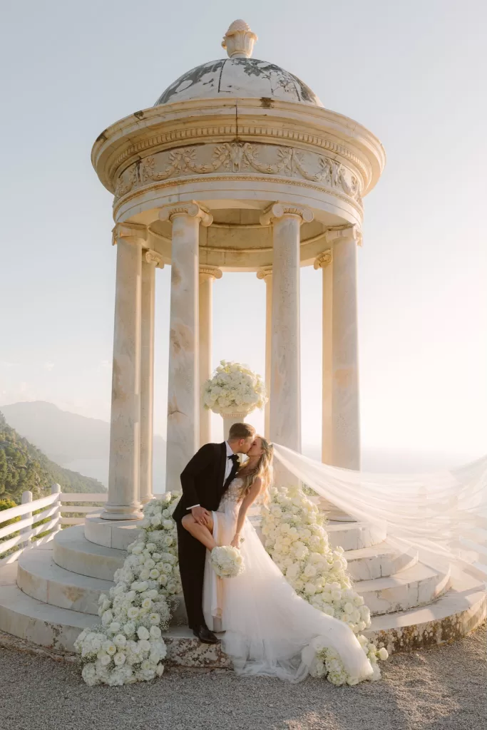 Romantic newlywed kiss beneath the marble pavilion at Son Marroig, framed by cascading white flowers and sea views.