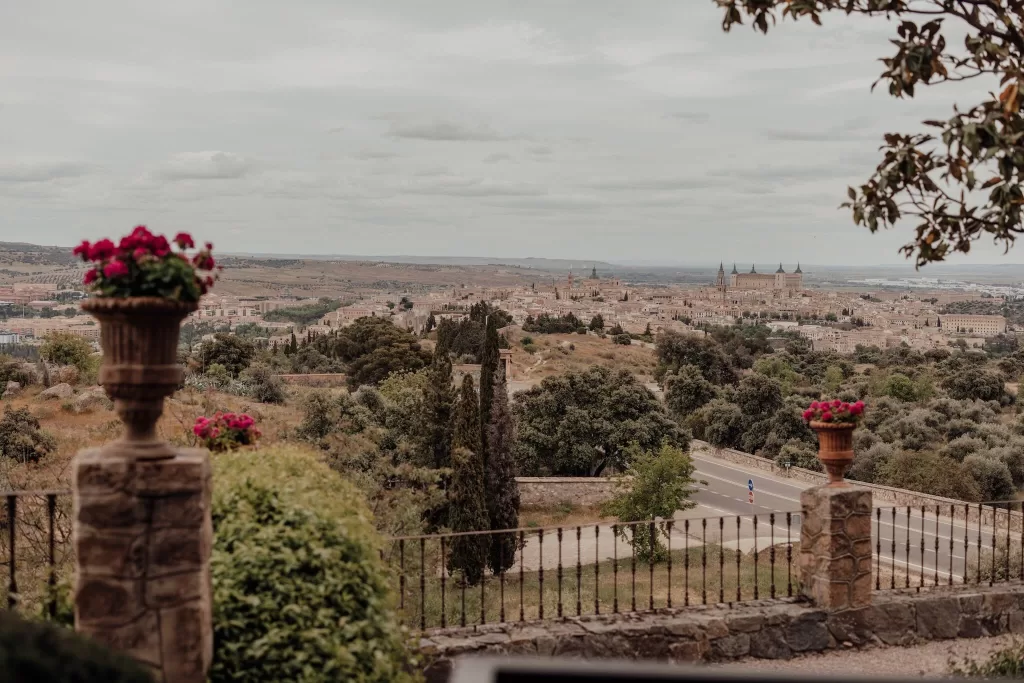 Expansive panoramic view of Toledo as seen from the gardens of Cigarral de la Mercedes, framed by terracotta planters and lush foliage.