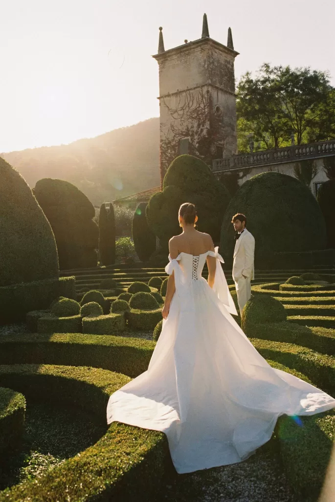 Bride walking through sculpted garden hedges in front of Villa Arvedi at sunset.