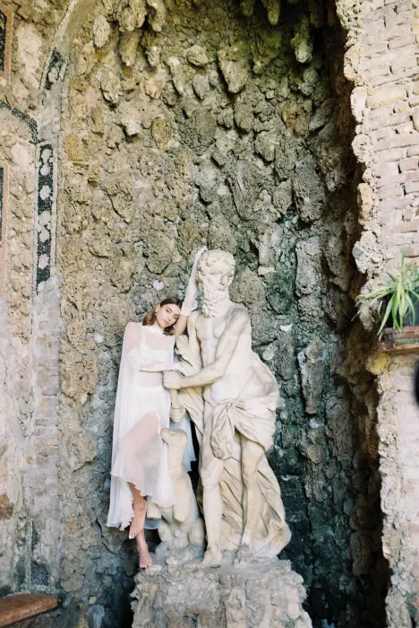 Bride in sheer robe leaning on a stone statue inside the grotto at Villa Arvedi.