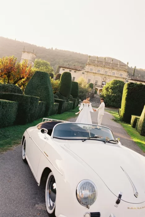 Bride and groom holding hands beside a classic white Porsche outside Villa Arvedi.