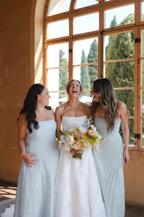 The bride laughing joyfully with her bridesmaids in soft blue dresses at Villa San Vigilio, framed by arched windows and cypress trees.