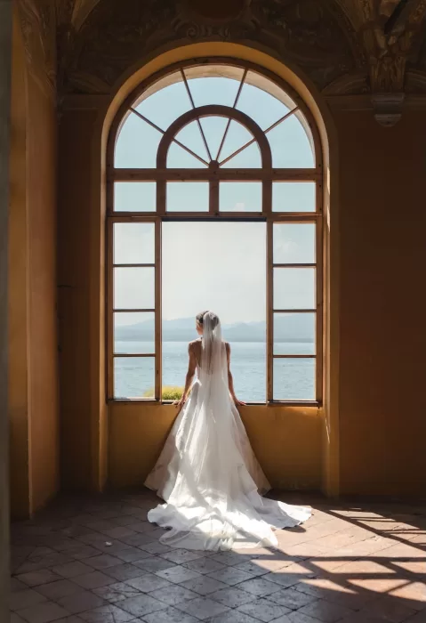 Bride gazes over Lake Garda from a grand arched window inside Villa San Vigilio, bathed in natural light.