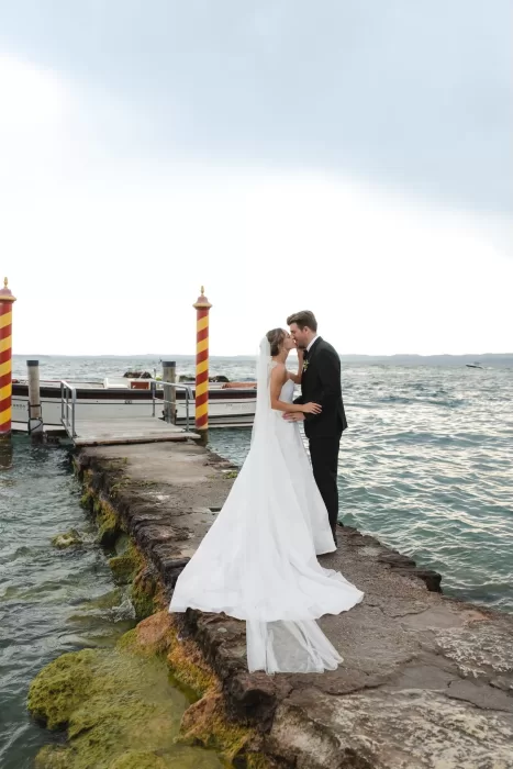 The couple shares a kiss at the end of a dock on Lake Garda, framed by striped pillars and water at Villa San Vigilio.