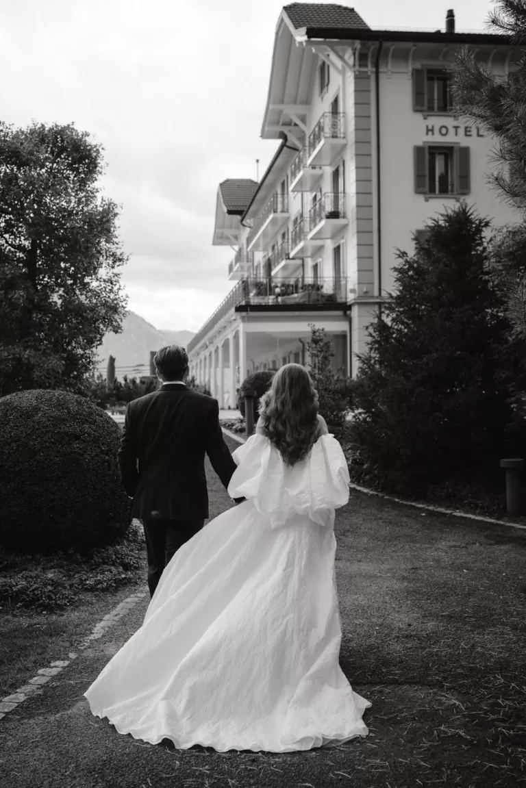 Bride and groom walk hand in hand away from Hotel Villa Honegg, their elegant attire trailing behind.