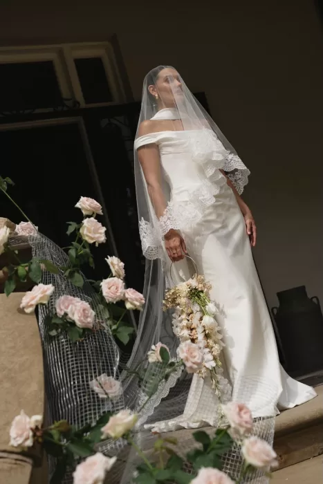 Elegant bride in lace-trimmed veil and cascading bouquet standing on the staircase adorned with roses at Wallalong House.