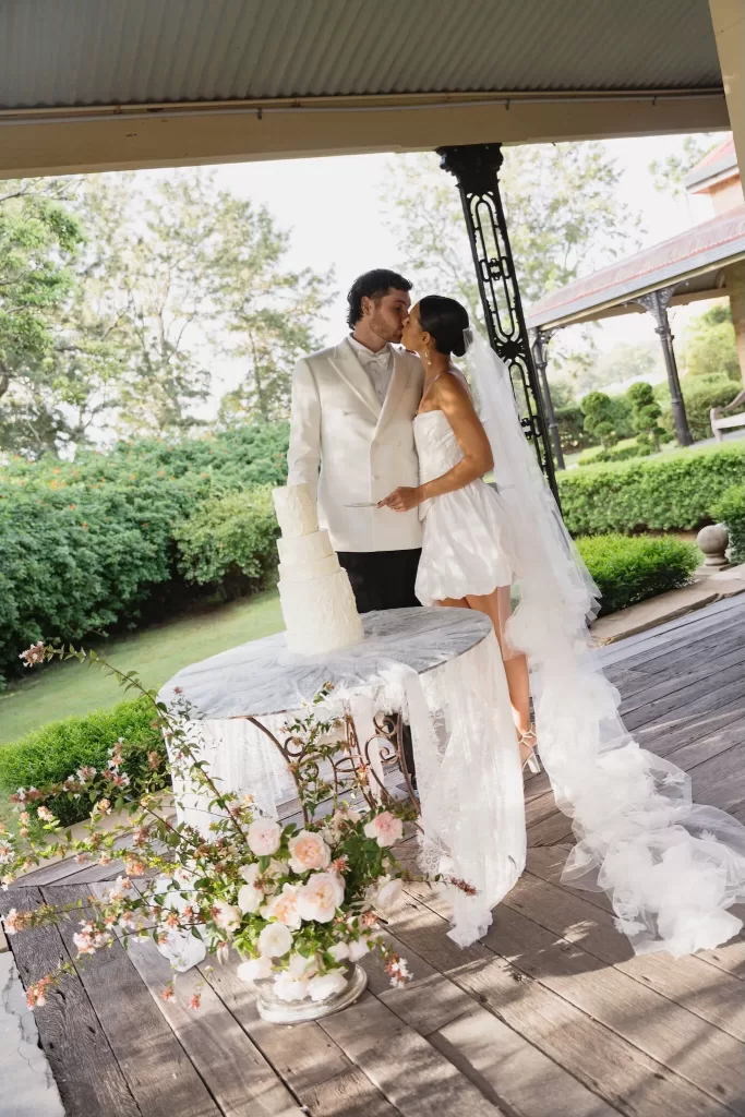 Bride and groom kissing beside their three-tier wedding cake on the veranda of Wallalong House.