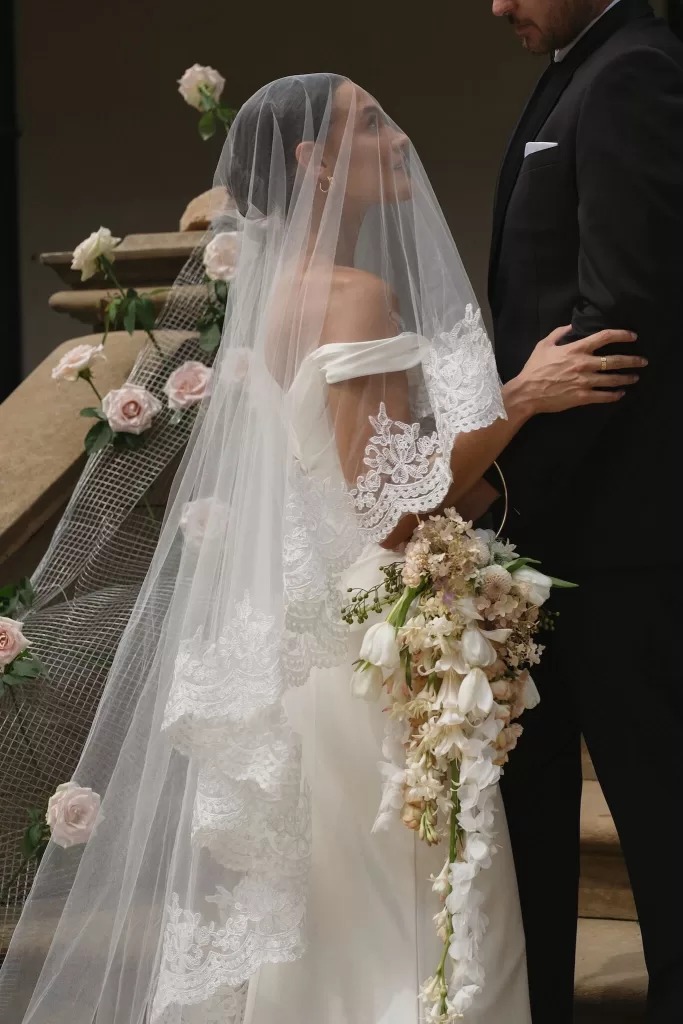 Bride in lace veil gazing at groom while holding cascading bouquet on the staircase of Wallalong House.