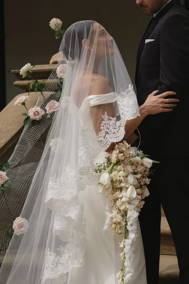 Bride in lace veil gazing at groom while holding cascading bouquet on the staircase of Wallalong House.