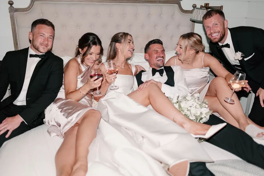 Bride and groom laughing on the bed with friends during celebrations at Middleton Lodge.