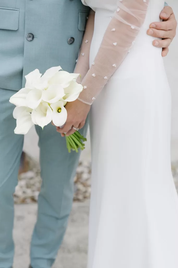 Close-up of bride holding white calla lilies beside groom in light blue suit at Winery Hans Igler.
