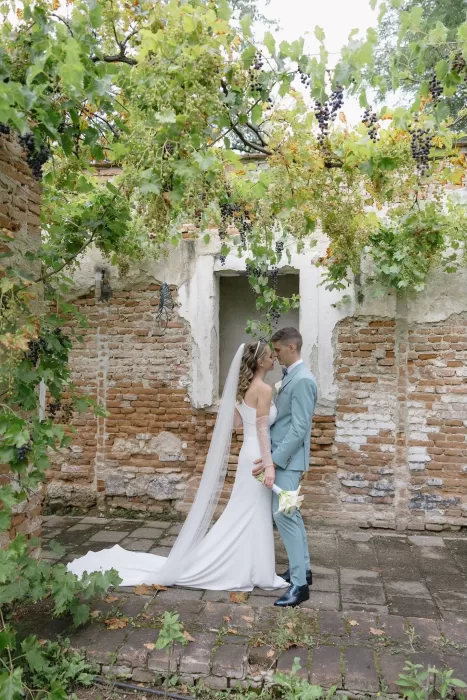 Bride and groom standing under hanging grapevines in rustic stone ruins at Winery Hans Igler.