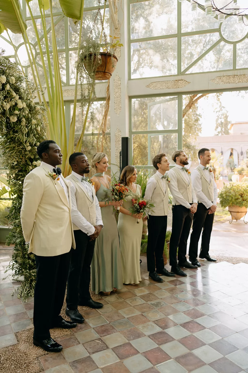 Wedding party lined up inside the sunlit glasshouse at Beldi Country Club, surrounded by lush greenery.