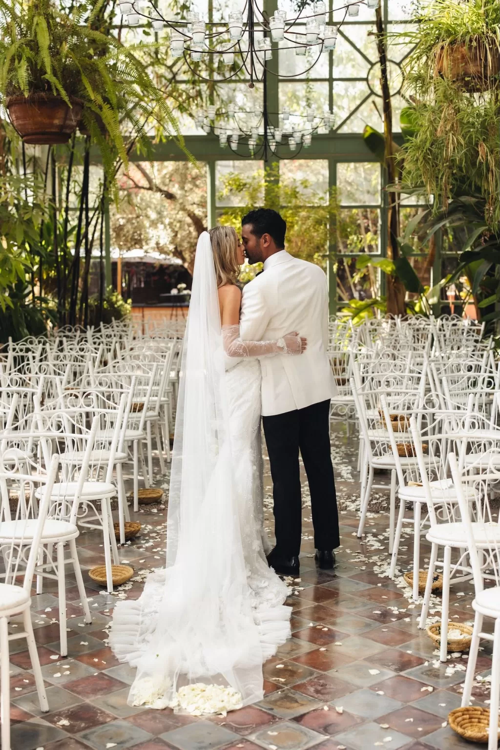 Newlyweds sharing a kiss inside the glasshouse at Beldi Country Club, framed by natural greenery and white chairs.