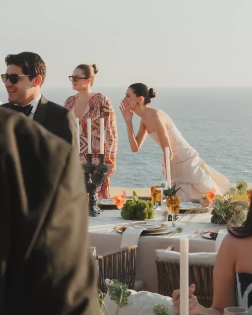 Bride laughing with guests by oceanfront reception table.