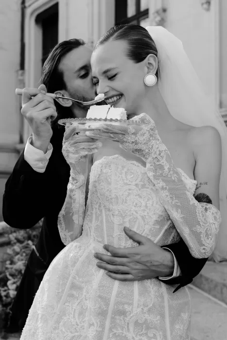 Groom feeding bride a slice of wedding cake as she laughs in front of Chateau Challain.