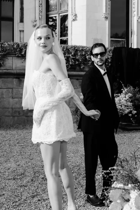 Bride in lace mini dress holding hands with groom in black tuxedo in Chateau Challain courtyard.