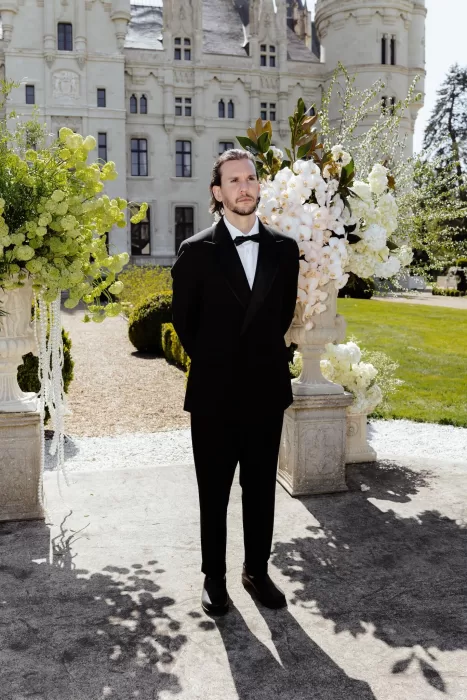 Groom in black tuxedo standing by floral arrangements with Chateau Challain as backdrop.
