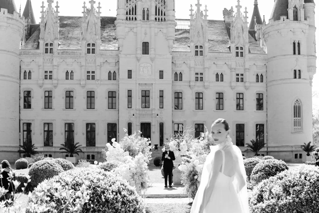 Bride in flowing gown walking through floral aisle in front of Chateau Challain’s grand facade.