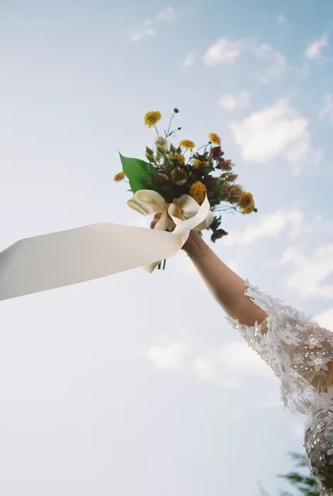 Bride raises bouquet of yellow roses with a trailing ribbon against a blue sky at House Boheme.