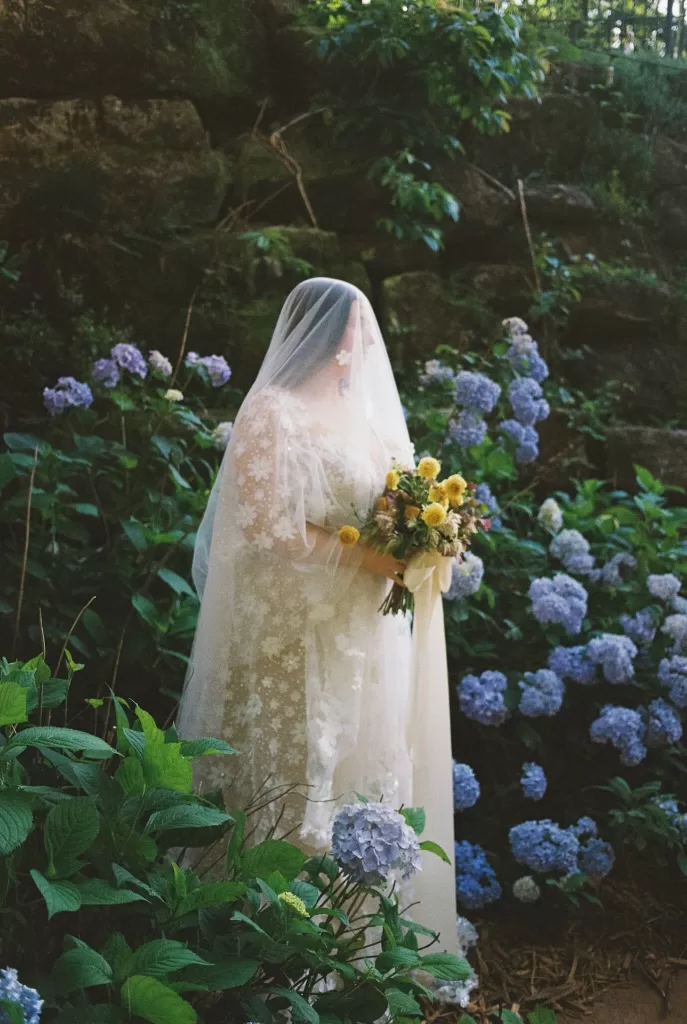 Bride in a floral lace gown and veil holding her bouquet among lush blue hydrangeas at House Boheme.