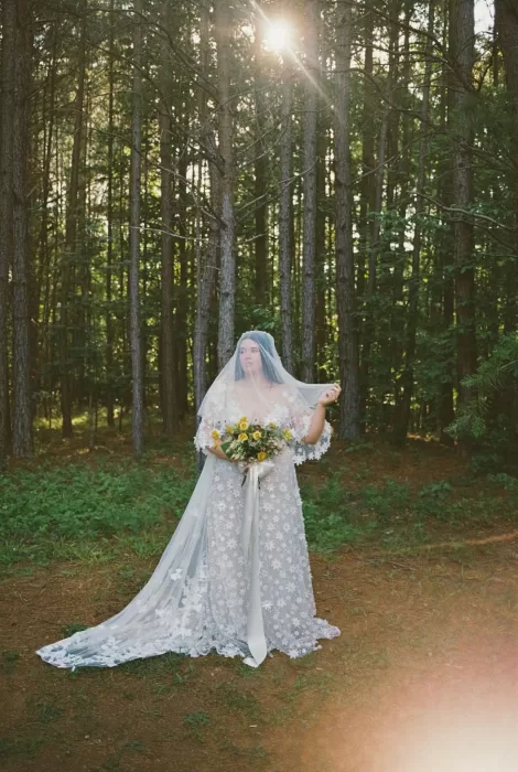 Bride in a floral lace gown and flowing veil holding yellow roses in the serene woodland at House Boheme