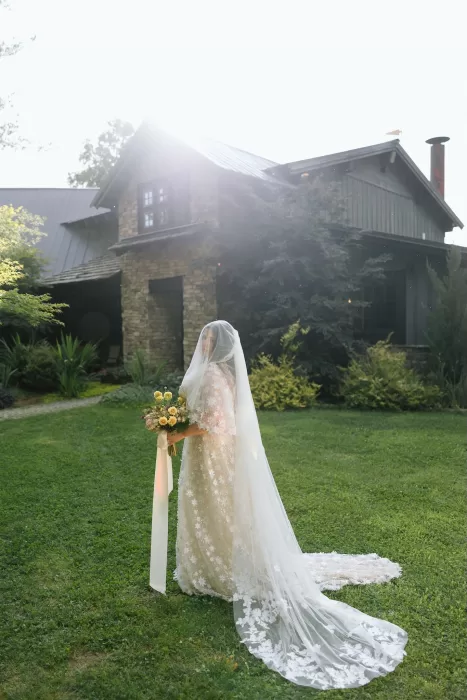 Bride in a long veil and floral gown stands in the garden with sunlight streaming over House Boheme.