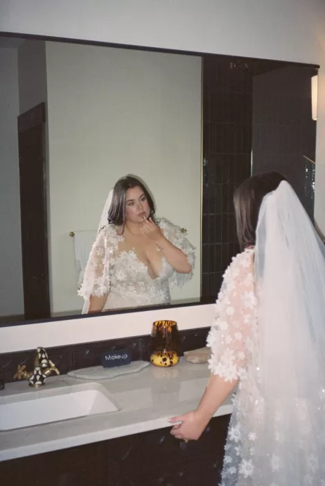 Bride touches up her makeup in the mirror, wearing a floral lace gown and veil at House Boheme.