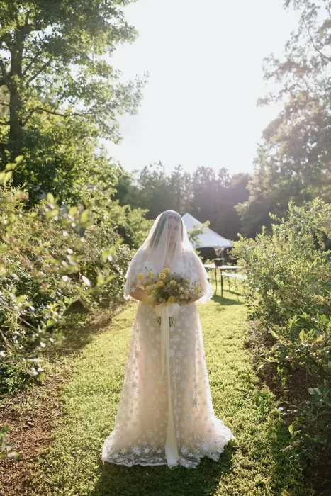 Bride in a long veil and lace gown holds a bouquet of yellow roses while standing in the orchard at House Boheme.