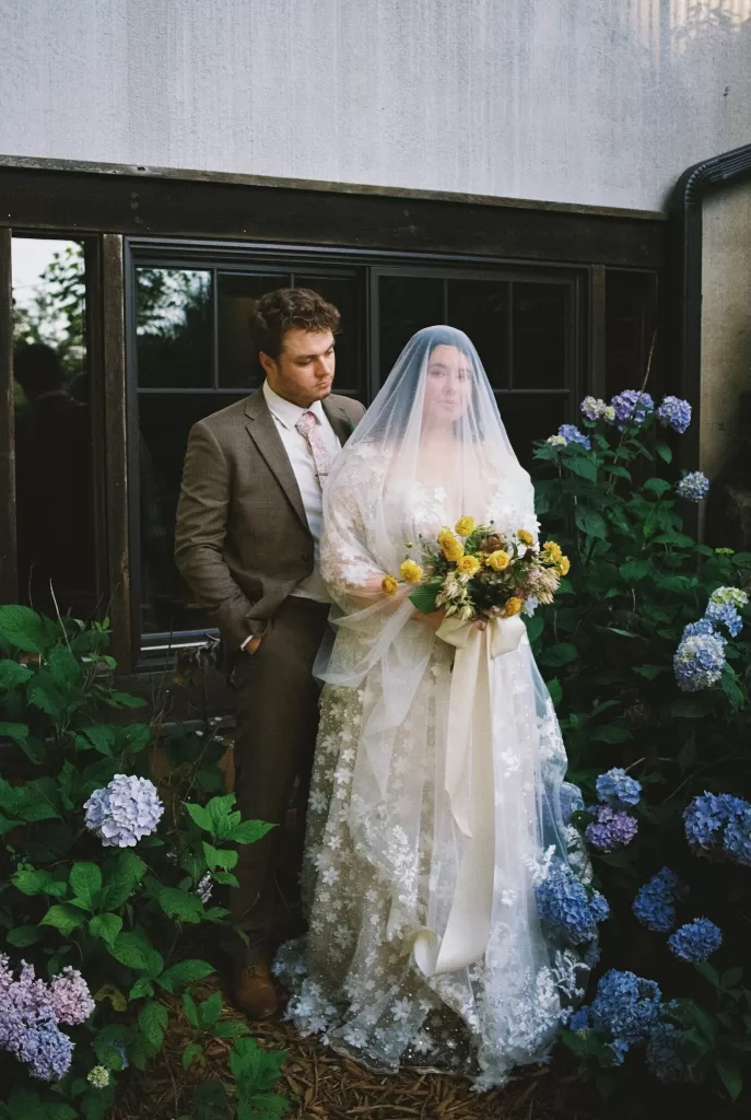 Romantic portrait of bride and groom by a window surrounded by blue hydrangeas at House Boheme.