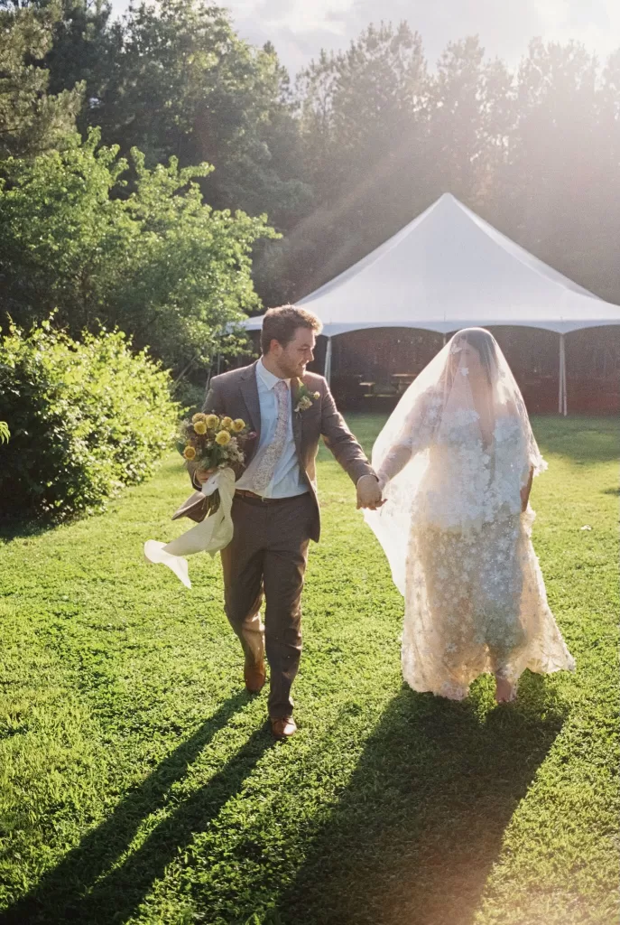 Bride and groom walk hand in hand across the lawn with a white wedding tent behind them at House Boheme.