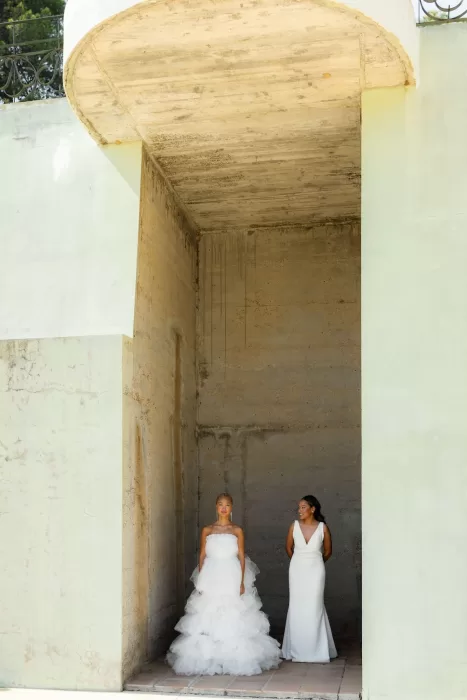 Two brides in elegant gowns standing against a minimalist concrete wall at La Finka 4.1.
