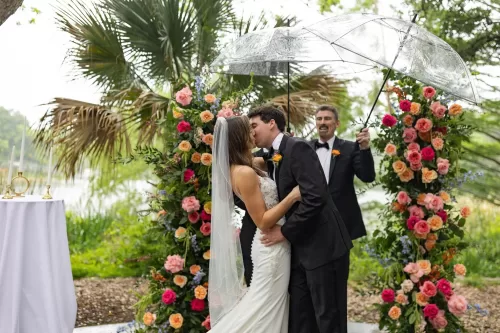 Newlyweds sharing a kiss beneath a floral arch and clear umbrella at Laguna Gloria.