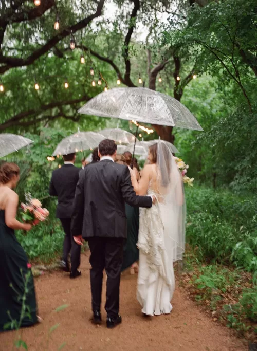 Wedding party walking with clear umbrellas through the trees at Laguna Gloria.