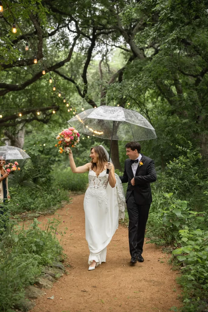 Bride and groom walking under umbrellas with string lights glowing at Laguna Gloria.