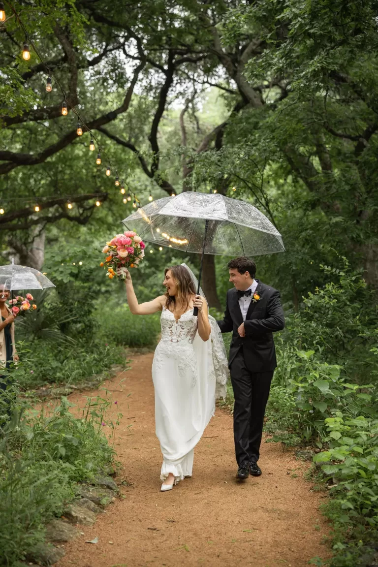 Bride and groom walking under umbrellas with string lights glowing at Laguna Gloria.