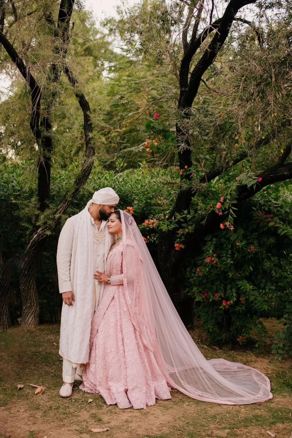 Bride and groom in traditional pink attire sharing a tender moment in lush garden, photographed by Lucky Elevens.