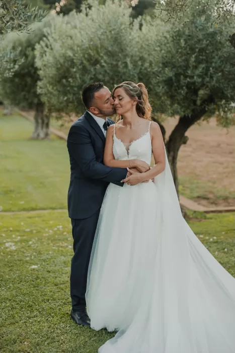 Groom kisses bride on the cheek during a romantic portrait in the gardens of Mas de Les Voltes.