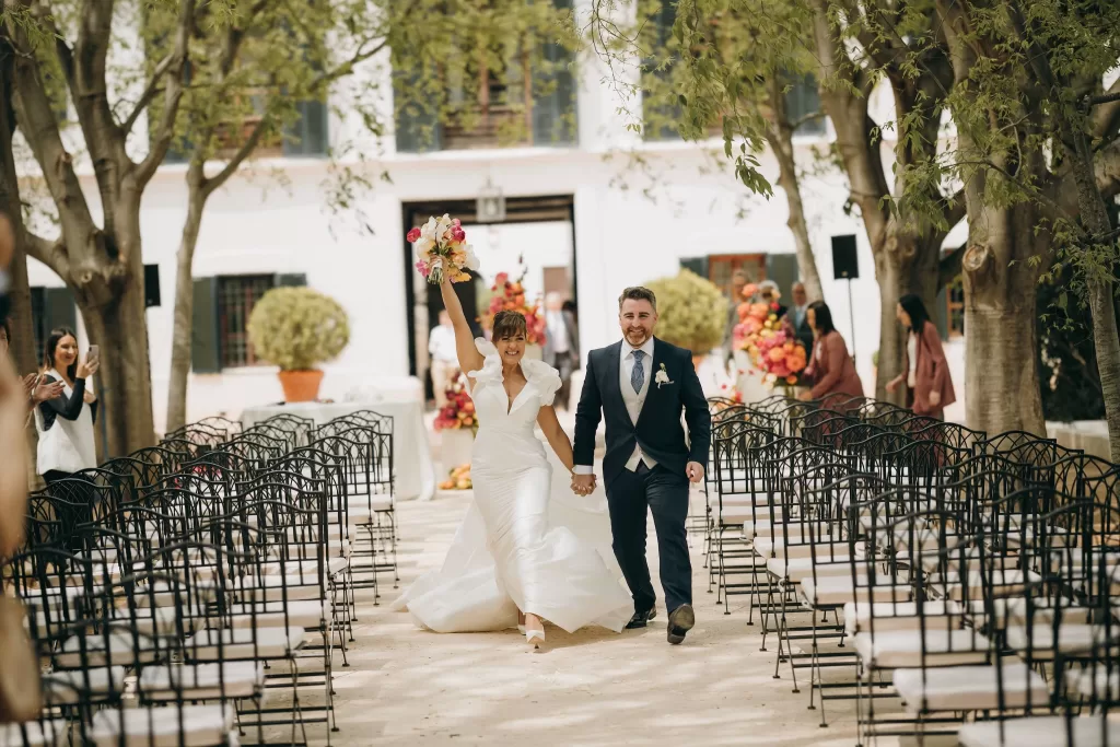 Newlyweds joyfully walk down the aisle at Masia Aldamar, bride raising bouquet in celebration.
