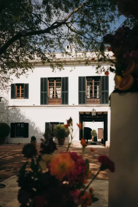 Framed view of Masia Aldamar’s white facade with green shutters and floral decor in the foreground.