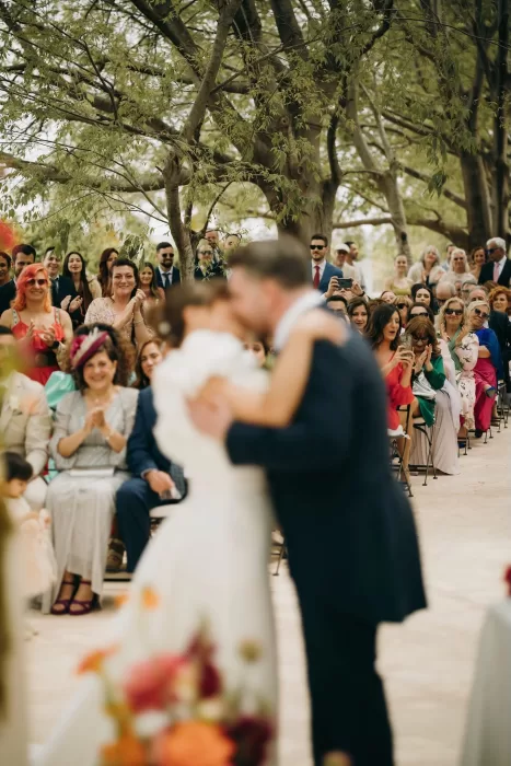Newlyweds share their first kiss surrounded by cheering guests under the trees at Masia Aldamar.