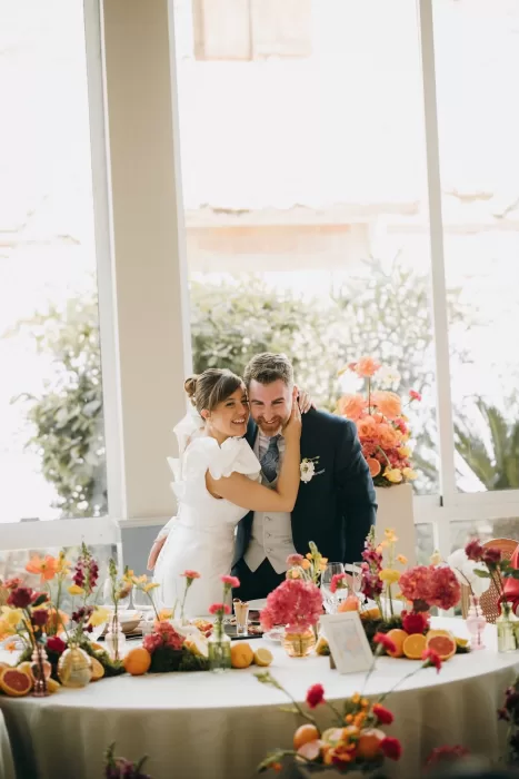 Bride embracing groom at a flower-filled reception table in Masia Aldamar.