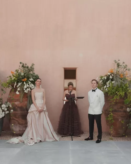 Wedding ceremony with couple standing before tall floral arrangements against a rustic wall.