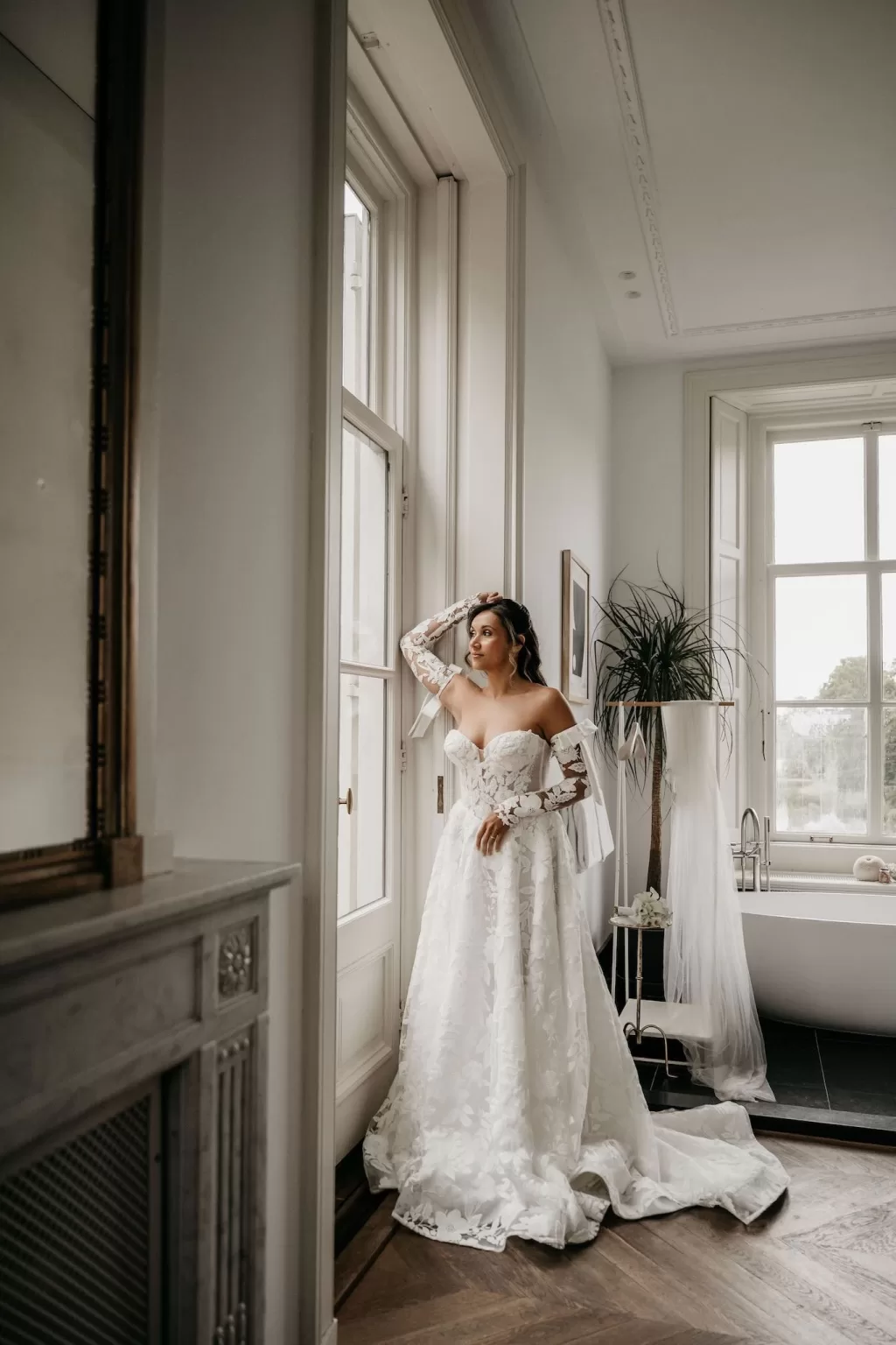Bride in lace gown standing by a window in a refined suite at Parc Broekhuizen.