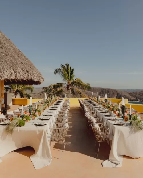 Long wedding reception tables decorated with flowers and candles on a terrace with palm trees.