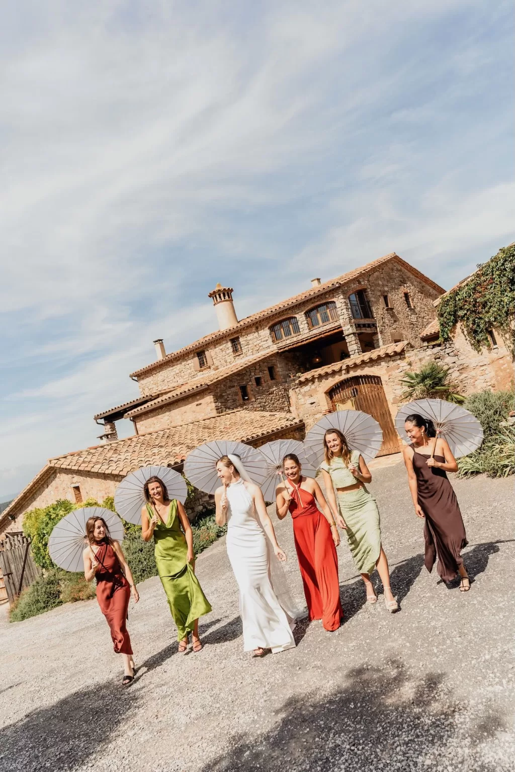 The bride and bridesmaids walk together under white parasols in front of the rustic stone building at This Must Be The Place.